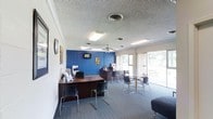 This single-floor commercial space is designed to accommodate both staff operations and client-facing activities. The layout is divided into distinct functional zones, beginning with a reception and work area anchored by a dark wood desk positioned near the entrance. Multiple chairs line the front of the desk, facilitating consultations or check-ins. Behind the desk, a multifunction printer and informational brochures are readily accessible.

A bold blue accent wall serves as a visual focal point at the rear of the space, complemented by a wall-mounted flat-screen television and decorative artwork. This design choice adds a distinctive contrast to the otherwise neutral tones of the room.

A dedicated seating area features a glass-topped table surrounded by modern chairs, providing a comfortable space for meetings or waiting. The space is illuminated by a combination of fluorescent ceiling fixtures and natural light streaming through large windows along one wall, offering views of the surrounding greenery outside. A ceiling fan aids in air circulation throughout the room. The textured ceiling and carpeted flooring contribute to the professional yet welcoming ambiance of the space.