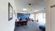 This single-floor commercial space is designed to accommodate both staff operations and client-facing activities. The layout is divided into distinct functional zones, beginning with a reception and work area anchored by a dark wood desk positioned near the entrance. Multiple chairs line the front of the desk, facilitating consultations or check-ins. Behind the desk, a multifunction printer and informational brochures are readily accessible.

A bold blue accent wall serves as a visual focal point at the rear of the space, complemented by a wall-mounted flat-screen television and decorative artwork. This design choice adds a distinctive contrast to the otherwise neutral tones of the room.

A dedicated seating area features a glass-topped table surrounded by modern chairs, providing a comfortable space for meetings or waiting. The space is illuminated by a combination of fluorescent ceiling fixtures and natural light streaming through large windows along one wall, offering views of the surrounding greenery outside. A ceiling fan aids in air circulation throughout the room. The textured ceiling and carpeted flooring contribute to the professional yet welcoming ambiance of the space.
