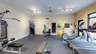 This fitness center offers a comprehensive workout environment thoughtfully arranged across a single floor. The space is finished with durable, dark carpet flooring suited for exercise use, and the walls are painted in a neutral, light tone that contributes to a clean and energizing atmosphere. Overhead, a combination of a ceiling fan and track lighting ensures the room remains well-lit and well-ventilated during use.

The layout accommodates a variety of training modalities. Strength training is supported by a multi-station cable machine and a dedicated leg press or extension machine, along with a flat weight bench for free weight exercises. Cardio equipment is also represented, with an elliptical trainer visible and additional machines positioned throughout the space.

Along one wall, a storage rack holds exercise accessories including stability balls in multiple sizes, a yoga mat, and resistance tools, keeping the space organized and versatile. Safety and instructional signage is posted on the wall, adding a practical, informative touch. HVAC vents are integrated into both the wall and ceiling, ensuring year-round climate comfort. Overall, the fitness center is a functional, well-maintained space designed to meet a broad range of exercise needs.