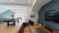 This single-floor amenity lounge is thoughtfully designed to accommodate both relaxed socializing and active recreation. The space is divided into two primary zones that flow seamlessly into one another, unified by light hardwood flooring and a bright, contemporary aesthetic.

The lounge area is anchored by a bold teal accent wall featuring a large wall-mounted flat-screen television, complemented by a tufted brown leather sofa, a navy accent chair, and a mid-century modern media console. The angled ceiling above this seating area adds architectural interest and an intimate atmosphere. Decorative shelving and carefully selected accessories contribute to the polished, curated feel of the lounge.

Adjacent to the lounge, the generously sized games room is bathed in natural light and equipped with a pool table, foosball table, and shuffleboard table. A stylish chandelier-style pendant light anchors the space, which also features a dartboard and ample open floor space for active play.

The kitchenette area is finished with a geometric patterned accent wall, light wood cabinetry, a white quartz countertop, stainless steel sink, coffee station, and under-counter refrigerator. Industrial-style pendant lights and bar stools complete this functional yet stylish refreshment zone. Double glass doors at the rear of the space provide natural light and outdoor access.