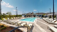 Located at 40000 Argento Way in Kannapolis, North Carolina, this single-level outdoor amenity area offers a well-appointed community retreat designed for relaxation and recreation. At its centerpiece is a generously sized lap pool with clearly marked lanes and crystal-clear water, surrounded by an expansive stone-paved deck lined with lounge chairs ideal for sunbathing. The pool area faces a charming clubhouse building with a light blue exterior, pitched roofline, and warm architectural detailing that anchors the space visually.

Adjacent to the pool, a covered pavilion serves as a true outdoor living room. Vaulted white-beamed ceilings and stone accent walls create a refined yet comfortable atmosphere, while pendant lighting and ceiling fans enhance the ambiance. A stone-clad fireplace with a mounted television provides a cozy gathering focal point, complemented by wicker seating with bold striped upholstery. The pavilion connects seamlessly to a fully equipped outdoor kitchen featuring a granite-topped stone island with bar seating, a built-in sink, and multiple stainless steel grills — perfect for entertaining.

Beyond the pavilion, a paved patio area with dining tables and chairs opens to manicured landscaping, mature trees, and views of the surrounding residential buildings. The entire space is enclosed by sleek black iron fencing, offering both security and an open, airy feel under the North Carolina sky.