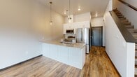 Located at 1021 Blazing Star Blvd in Altoona, Wisconsin, this two-bedroom, two-bathroom home spans two floors with a well-designed layout. The main level features an open-concept kitchen and living area highlighted by rich wood-look plank flooring, recessed lighting, and abundant natural light through sliding glass doors that open to outdoor views of the surrounding greenery. The kitchen is equipped with white shaker cabinetry, stainless steel appliances including a refrigerator, microwave, and range, complemented by a granite-topped center island with pendant lighting above — a standout feature of the main living space. The living room is bright and spacious, with additional sliding door access to the outdoors. Upstairs, the generously sized primary bedroom features plush carpeting, a ceiling fan, and a private en-suite bathroom and walk-in closet with built-in shelving for ample storage. The second bedroom is well-lit with multiple windows and a stylish barn-style door. Bathrooms are finished with granite vanity tops and white cabinetry. A convenient mudroom area with bench seating and coat hooks adds everyday practicality. Dark wood trim and door accents throughout provide a cohesive, modern aesthetic across both levels.