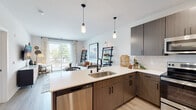 This single-floor residence showcases a thoughtfully designed open-concept layout that seamlessly connects the kitchen, dining, and living areas. The kitchen is outfitted with dark wood-grain flat-front cabinetry, quartz countertops, a subway tile backsplash, and a full suite of stainless steel appliances, including a range, built-in microwave, refrigerator, and dishwasher. A peninsula with an undermount sink serves as both a functional workspace and a natural divider between the kitchen and living area, illuminated by two sleek pendant lights overhead.

The living area is bright and welcoming, with light wood-style flooring and large sliding glass doors that open to a private balcony, flooding the space with natural light and providing outdoor views. The two bedrooms are carpeted and well-proportioned, each featuring ample closet space, large windows, and ceiling fans for year-round comfort. The bathroom includes a quartz vanity countertop, an undermount sink, a large mirror with a bold frame, and a shower-tub combination. Throughout the residence, neutral tones and modern finishes create a cohesive and contemporary aesthetic.