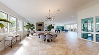 This expansive single-floor community clubhouse offers a generous, open-concept floor plan designed for both relaxation and recreation. The space is unified by large-format travertine-style tile flooring that flows seamlessly throughout, complementing the bright white walls and vaulted ceilings that amplify the sense of openness.

The living and dining areas serve as the heart of the space, anchored by a striking stacked-stone fireplace with a wall-mounted television above. A round wood dining table surrounded by contemporary upholstered chairs sits beneath a dramatic sputnik-style chandelier, creating an inviting gathering spot. Lush potted tropical plants are positioned throughout, adding warmth and life to the interior.

The recreation zone features a full-size pool table and a foosball table, while a vibrant teal-accented computer station provides a dedicated business center area. A row of mid-century modern seating lines a window wall overlooking lush, verdant landscaping, flooding the space with natural light and outdoor views.

Adjacent to the main area, a private office with a large executive desk connects to a glass-enclosed fitness center equipped with exercise machines. French doors and expansive windows on multiple walls ensure the entire space remains bright, airy, and connected to the surrounding greenery.