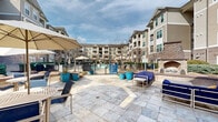 Located at 7880 Triangle Promenade Dr in Raleigh, NC, this outdoor amenity area is the social centerpiece of a multi-building residential community. The space is designed with a resort-style aesthetic, anchored by a large swimming pool enclosed within a wrought-iron fence and surrounded by an expansive stone-tiled pool deck. The deck is furnished with lounge chairs, market umbrellas, and comfortable seating arrangements, offering ample space for relaxation and recreation under open skies.

Adjacent to the pool area, a covered pergola structure provides a shaded outdoor gathering space equipped with a grilling station and dining seating—ideal for outdoor entertaining. Nearby, additional lounge chairs are arranged along a concrete sun deck, framed by manicured landscaping beds featuring ornamental shrubs, junipers, and seasonal plantings.

Curving concrete walkways wind throughout the grounds, connecting the various amenity zones and building entries. The building exteriors feature a classic combination of brick and neutral-toned siding, with arched entryways, covered breezeway corridors lit by wall-mounted lanterns, and private balconies on upper floors. The thoughtfully maintained landscaping with mulched beds, trimmed hedgerows, and mature trees enhances the community's welcoming, park-like atmosphere throughout.