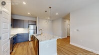 This contemporary single-floor residence in Wake Forest, NC offers a thoughtfully designed layout with modern finishes throughout. The open-concept kitchen serves as the heart of the home, anchored by a spacious granite-topped island with an undermount sink and brushed gold faucet. Two elegant glass pendant lights hang above the island, complementing the two-tone cabinetry — sleek gray upper cabinets paired with navy-blue lower cabinets — and a marble-style tile backsplash. A full suite of stainless steel appliances, including a refrigerator, range, microwave, and dishwasher, completes the kitchen. Warm wood-style flooring flows seamlessly throughout the entire unit, creating a cohesive and inviting atmosphere. The living area benefits from natural light streaming through a glass door with a transom window above, leading to an exterior access point. The bedrooms are bright and well-proportioned, with large windows fitted with blinds and ceiling fans. One bedroom includes a dedicated walk-in closet with wire shelving. The bathroom features a granite-topped vanity with gray cabinetry, wood-style flooring, and a shower-tub combination. A dedicated laundry area houses a full-size top-loading washer and dryer with overhead wire shelving for added storage convenience.