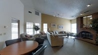 This single-floor communal space offers a comfortable and functional layout designed for relaxed gathering and conversation. The room is distinguished by its vaulted ceiling, which lends a sense of openness and airiness to the interior. Textured ceilings and warm tan-toned accent walls contribute to the inviting atmosphere, complemented by neutral-colored carpeting that runs throughout the space.

The lounge area is generously furnished with multiple seating arrangements, including a large sofa and loveseat combination paired with coordinating armchairs, all upholstered in earthy tones. Decorative throw pillows add a touch of color and comfort to the seating. A glass-top coffee table anchors the central seating area, while additional armchairs are positioned near the windows to take advantage of the natural light.

Track lighting installed along the ceiling provides ambient illumination, supplemented by a floor lamp near the sofa grouping. Several framed artworks adorn the tan feature wall, adding visual interest. A large potted tree near the windows introduces a natural element to the space. In the foreground, a round dining table with chairs provides an additional functional area for seated use.