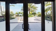 This single-floor space is designed with a clean, modern aesthetic that prioritizes openness and functionality. The interior is characterized by dramatically high ceilings that amplify the sense of volume, crisp white walls, and large-format neutral-toned tile flooring accented by dark baseboards that provide sharp visual contrast throughout.

The main open area is generously proportioned, offering flexible use potential. Architectural niches and angled wall cuts add visual interest and dimension to the otherwise minimalist interior. A wall-mounted dispenser station and integrated smart panels suggest the space is configured for shared or amenity use. Framed artwork provides a focal point along one wall, adding a refined touch to the open layout.

Floor-to-ceiling glass doors and sliding glass panels connect the interior directly to an expansive outdoor terrace, flooding the space with natural light. The terrace is beautifully landscaped with tall palm trees, tropical plantings, and contemporary white planters, adjacent to what appears to be a pool area. Views of the surrounding urban skyline and blue skies reinforce the bright, resort-inspired ambiance. The seamless indoor-outdoor connection is a defining feature of this well-appointed space.