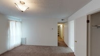 This single-floor residential unit offers a practical and straightforward layout designed for comfortable everyday living. The living area is carpeted and filled with natural light from large windows dressed with sheer curtains, complemented by a classic flush-mount light fixture and a textured popcorn ceiling. A wall-mounted thermostat and baseboard heating units are present throughout, ensuring climate control in each room.

The kitchen is efficiently designed with white cabinetry offering generous upper and lower storage, white laminate countertops, and a full suite of white appliances including a coil-burner electric range, overhead range hood, refrigerator, and dishwasher. Light vinyl flooring runs through the kitchen, which connects directly to the living areas via a wide doorway.

The bedrooms are carpeted and well-sized, each featuring textured ceilings and sliding or hinged closet doors with built-in hanging rods for storage. One bedroom includes access to a walk-in style closet area with open shelving.

Both bathrooms are finished with neutral tones and feature tub-shower combinations with panel surrounds, tile flooring, and granite-topped vanities. A dedicated bonus room with light carpet and a horizontal window provides flexible additional space. The overall aesthetic is clean, neutral, and move-in ready.