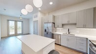 Located at 8157 Ederville Rd in Fort Worth, TX, this one-bedroom, one-bathroom unit offers a clean, modern design with a cohesive neutral palette throughout. The open-concept living and kitchen area is anchored by a sleek white island beneath elegant pendant lighting, with warm wood-look flooring extending throughout the main living spaces. The kitchen is well-equipped with gray shaker-style cabinetry, white quartz countertops, a subway tile backsplash, and a full suite of stainless steel appliances, including a French door refrigerator, dishwasher, microwave, and range. The living area features recessed lighting and direct access to a private balcony through double glass doors, complemented by a ceiling fan overhead. The bedroom is a comfortable, carpeted retreat with a large window framing outdoor views and filling the space with natural light. The bathroom is finished with light wood-look flooring, gray vanity cabinetry, white countertops, and provides convenient access to a dedicated walk-in closet with built-in shelving. Throughout the unit, tall ceilings and neutral tones create a bright, airy ambiance that feels open and inviting.