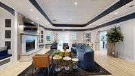 The Fonthill Court Clubhouse Living Room in Langhorne, PA is a thoughtfully designed community gathering space that blends modern style with comfortable functionality. The main lounge anchors the space with a distinctive tray ceiling accented in deep navy, recessed lighting, and light hardwood flooring throughout. A marble-surround fireplace with a sleek wood-slat frame and wall-mounted TV serves as the room's focal point, flanked by built-in arched shelving with a cozy window seat. Curved navy sofas, leather armchairs, and gold-toned coffee tables complete the inviting seating arrangement. An adjacent sunlit dining and lounge area features vaulted ceilings and oversized windows that flood the space with natural light and offer views of the surrounding greenery. Multiple seating configurations — including a round wood dining table, casual café-style tables, and bench seating — offer flexible use for residents. The fully equipped kitchen showcases white shaker cabinetry with gold hardware, quartz countertops, a geometric blue-and-white tile backsplash, stainless steel appliances, and a butcher block island with bar seating. A private meeting room with a marble-top conference table and sliding barn door provides a quiet space for gatherings or work. Light-toned hardwood flooring and soft grey walls unify the entire clubhouse with a clean, contemporary aesthetic.