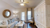 Located at 2030 Wildts Battery Blvd on the charming Johns Island in South Carolina, this single-story home blends coastal warmth with classic Southern character. The living area immediately sets the tone with rich, dark hardwood flooring that contrasts beautifully against the neutral, warm-toned walls. Natural light pours in through white-shuttered windows dressed with elegant patterned drapery in soft blues and grays, lending the space a relaxed, coastal aesthetic. A ceiling fan with a built-in light fixture hangs overhead, adding both functionality and a casual, inviting feel to the room. The living space is thoughtfully appointed, featuring a comfortable slipcovered sofa, a rustic whitewashed coffee table, and a classic accent chair with coordinating patterned upholstery. A large, distressed white armoire anchors one side of the room, offering both storage and a refined farmhouse touch. Decorative lighting and carefully chosen accessories throughout reflect a cohesive, beachy-chic style. A glass-paned door leads to the outdoors, further enhancing the sense of light and connection to the natural surroundings. Overall, this home offers a serene, well-curated retreat with a welcoming ambiance suited to coastal low-country living.
