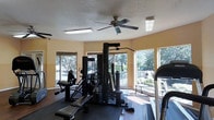 This fitness center is designed to provide a functional and comfortable workout environment. The space is outfitted with a multi-station cable and weight machine that accommodates a variety of strength training exercises, along with cardio equipment including a treadmill. Rubber flooring in the equipment area offers safety and stability during workouts. A textured ceiling with a ceiling fan and flush-mounted fluorescent light fixtures ensures the space remains well-lit and properly ventilated throughout the day. Large windows wrap around the room, flooding the interior with natural light and offering pleasant views of the surrounding landscaping and outdoor amenity areas, including what appears to be a pool or courtyard. The warm, neutral-toned walls contribute to an inviting and energizing atmosphere. The layout is open and organized, allowing users to move freely between equipment stations. Overall, this fitness center delivers a practical and welcoming setting suited for daily exercise routines.