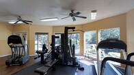 This fitness center is designed to provide a functional and comfortable workout environment. The space is outfitted with a multi-station cable and weight machine that accommodates a variety of strength training exercises, along with cardio equipment including a treadmill. Rubber flooring in the equipment area offers safety and stability during workouts. A textured ceiling with a ceiling fan and flush-mounted fluorescent light fixtures ensures the space remains well-lit and properly ventilated throughout the day. Large windows wrap around the room, flooding the interior with natural light and offering pleasant views of the surrounding landscaping and outdoor amenity areas, including what appears to be a pool or courtyard. The warm, neutral-toned walls contribute to an inviting and energizing atmosphere. The layout is open and organized, allowing users to move freely between equipment stations. Overall, this fitness center delivers a practical and welcoming setting suited for daily exercise routines.