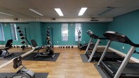 This dedicated fitness room offers a practical and functional space for exercise and wellness. The room features warm hardwood-style flooring complemented by bold teal accent walls, creating an energizing atmosphere for workouts. Overhead fluorescent lighting ensures the space is well-lit throughout, while windows along the back wall allow for natural light and outdoor views, adding an open, refreshing feel to the environment.

The room is equipped with a variety of cardio machines, including a treadmill, an elliptical trainer, and a stationary recumbent bike, each positioned on protective rubber mats to preserve the flooring. A set of kettlebells of varying weights is neatly arranged along the wall, providing options for strength and functional training. A wall-mounted medicine ball rack adds additional versatility for dynamic exercises and conditioning work.

The layout is thoughtfully organized to allow comfortable movement between stations, making it easy to transition between different types of workouts. A drop ceiling with recessed panel lighting keeps the space feeling clean and tidy. Overall, this fitness room is a comprehensive, well-maintained facility suited for cardiovascular training, strength building, and general fitness.