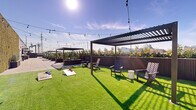 Located at 821 E Main St in Stamford, Connecticut, this rooftop amenity deck offers a thoughtfully designed outdoor retreat with a variety of recreational and social spaces. The expansive rooftop is divided into distinct zones, each catering to a different experience while maintaining a cohesive, modern aesthetic throughout.

A generous artificial turf lawn serves as the centerpiece of the rooftop, lined with Adirondack-style seating and multiple sleek pergolas draped with café string lights that create an inviting ambiance both day and night. Cornhole boards are set up across the lawn, making it an ideal space for casual outdoor recreation and social gatherings.

Adjacent to the turf area, a dedicated children's play zone features a charming playhouse, stepping stones, a Connect Four game, and additional play structures, all set against a living green wall backdrop and open sky views of the surrounding Stamford skyline.

A paved terrace section provides a more structured setting with outdoor dining tables and chairs, surrounded by tall planted greenery in modern wood-paneled planters that add texture and privacy. The planting throughout the rooftop softens the urban environment and enhances the sense of a garden-like escape.

The entire deck enjoys abundant natural sunlight and panoramic views of the Stamford cityscape, making it a versatile amenity space suited for relaxation, entertainment, and community engagement.