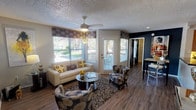 This single-floor space centers around a well-appointed living area designed with both comfort and style in mind. The room features warm wood-look hardwood flooring that runs throughout, anchored by a textured area rug that defines the seating arrangement. A cream sofa adorned with bold sunflower-patterned throw pillows pairs with two coordinating patterned accent chairs, creating an inviting and cohesive aesthetic.

Large windows along the front wall, dressed with decorative patterned valances and horizontal blinds, flood the space with natural light while offering views of a landscaped outdoor area. A glass-paneled door provides direct outdoor access and adds to the bright, airy atmosphere. A ceiling fan with a light fixture is centrally positioned overhead, complementing the textured popcorn ceiling.

A round glass-top coffee table serves as a focal point within the seating area, while recessed lighting enhances the overall illumination. Adjacent to the living area, a glimpse of a dining space is visible, featuring a high-top table and bar stools against a deep navy accent wall, offering a striking contrast to the lighter living zone. The layout is open and functional, balancing comfort with a modern decorative sensibility.