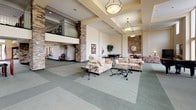 Located at 650 Woodward St in San Marcos, California, this single-story community clubhouse is designed to serve as a welcoming hub for social and recreational activities. The centerpiece of the facility is a grand, open lounge area distinguished by soaring double-height ceilings, elegant pendant lighting, and stacked stone accent columns that lend a warm, upscale character to the space. Comfortable seating arrangements, including floral sofas and glass-topped coffee tables, invite relaxation, while a billiards table adds a recreational element. A mezzanine-level overlook with metal railings adds architectural depth to the main hall. Floor-to-ceiling windows and double glass doors flood the lounge with natural light and offer direct views of — and access to — the outdoor pool and patio area, complete with shaded umbrellas and seating. A baby grand piano positioned near the pool entrance further enhances the refined ambiance. The clubhouse also features a fully equipped kitchen with white cabinetry, granite-style countertops, a center island, and white appliances, complemented by a pass-through bar with counter seating. An adjacent casual dining area with tile flooring and bistro-style seating provides a relaxed space for informal meals or meetings. Throughout, the design balances elegance and functionality, making this clubhouse a versatile venue for community events and everyday enjoyment.