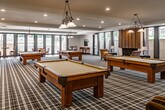 Located at 190 City Blvd W in Orange, California, this single-floor community amenity space is thoughtfully designed for recreation and socializing. The centerpiece of the room is a collection of full-size billiard tables, each illuminated by elegant wrought-iron pendant light fixtures that add a classic, refined touch to the atmosphere. The floors are covered in a bold black, white, and gold plaid carpet that lends a distinctive, club-like character to the space throughout.

Recessed ceiling lighting complements the pendant fixtures, ensuring the room is well-lit across all areas. A cozy lounge corner anchors one end of the space, featuring a wood-paneled fireplace flanked by armchairs, offering a relaxed retreat from the activity of the game area. On the opposite end, comfortable sofas and a wall-mounted flat-screen television create an inviting secondary seating area.

Large sliding glass doors and floor-to-ceiling windows along the perimeter flood the interior with natural light and provide direct access to an outdoor patio, seamlessly blending indoor and outdoor living. An adjacent corridor with decorative iron railings and staircase access connects the game room to additional amenities, including what appears to be a fitness area. The overall design is polished and functional, suited for an active community lifestyle.