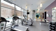 This single-floor fitness center is thoughtfully designed to provide a bright, motivating environment for exercise. Floor-to-ceiling windows line one side of the space, flooding the room with natural light and offering views of the surrounding outdoor area, including what appears to be a pool terrace. The cardio zone is well-equipped with treadmills, elliptical trainers, and a stationary bike, all arranged to take advantage of the expansive window views. Pendant lighting hangs from the white drop ceiling, adding a clean, modern aesthetic to the space. A wall-mounted flat-screen television is positioned centrally for entertainment during workouts. One wall features large glass panels and mirrors, which visually expand the space and enhance the open, airy ambiance. A medicine ball rack with an assortment of weighted balls is visible along the mirrored wall, adding to the functional fitness offerings. Beyond the glass partition, a lounge area is visible, furnished with contemporary yellow seating and colorful accents, providing a welcoming space to relax before or after a workout. The durable rubber flooring throughout ensures safety and practicality. Overall, the space strikes a balance between modern design and functional fitness amenities.