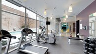 This single-floor fitness center is thoughtfully designed to provide a bright, motivating environment for exercise. Floor-to-ceiling windows line one side of the space, flooding the room with natural light and offering views of the surrounding outdoor area, including what appears to be a pool terrace. The cardio zone is well-equipped with treadmills, elliptical trainers, and a stationary bike, all arranged to take advantage of the expansive window views. Pendant lighting hangs from the white drop ceiling, adding a clean, modern aesthetic to the space. A wall-mounted flat-screen television is positioned centrally for entertainment during workouts. One wall features large glass panels and mirrors, which visually expand the space and enhance the open, airy ambiance. A medicine ball rack with an assortment of weighted balls is visible along the mirrored wall, adding to the functional fitness offerings. Beyond the glass partition, a lounge area is visible, furnished with contemporary yellow seating and colorful accents, providing a welcoming space to relax before or after a workout. The durable rubber flooring throughout ensures safety and practicality. Overall, the space strikes a balance between modern design and functional fitness amenities.