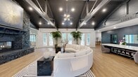 Located at 9727 Touchton Rd in Jacksonville, Florida, this single-floor clubhouse space delivers an impressive blend of industrial and contemporary design. The centerpiece of the main lounge is a floor-to-ceiling stacked stone fireplace, anchoring a spacious open area with soaring vaulted ceilings accented by dark-painted exposed steel beams and trusses. A striking Sputnik chandelier hangs above a curved, cream sectional sofa paired with black marble side tables and a geometric-patterned area rug, creating a sophisticated and inviting gathering space. Large windows and glass doors line the perimeter, flooding the interior with natural light and offering views of the surrounding greenery. Light wood-look flooring transitions to polished concrete in other areas, adding textural contrast throughout. A lounge area near the bar features deep navy shiplap walls, a white marble-topped bar with sleek black stools, and pendant globe lighting, lending a moody, upscale ambiance. A bright, windowed dining area with plantation shutters provides a quieter setting ideal for casual meetings or social gatherings. A dedicated conference room with a live-edge wood table, blue task chairs, a wall-mounted display, and designer wallcovering completes the functional offerings of this well-appointed community space.