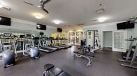 This single-floor fitness center offers a comprehensive workout environment designed to accommodate a wide range of exercise routines. The space is finished with soft blue-gray walls and durable rubber flooring, creating a clean and functional atmosphere throughout.

The cardio area is prominently featured, housing a row of treadmills and an elliptical trainer aligned along one wall. Multiple flat-screen TVs are mounted at eye level, providing entertainment during workouts. Ceiling fans are installed throughout, contributing to air circulation and comfort during use.

The strength training section includes a multi-station cable machine, a weight bench, and a well-stocked dumbbell rack with weights arranged in ascending order. Full-length mirrors line portions of the walls, allowing users to monitor form and enhancing the sense of openness within the space.

Natural light is a defining feature, with large multi-pane windows along exterior walls offering views of surrounding greenery. French doors with glass panels serve as the primary entrance, further connecting the interior to the outdoors and allowing additional light to filter in.

Decorative flush-mount light fixtures complement the classic architectural detailing, including crown molding, reinforcing the polished and well-maintained character of this fitness facility.