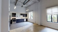 Unit 201 is a 1-bedroom, 1-bathroom residence that showcases a striking industrial-modern aesthetic across a single level. The open-concept layout is anchored by soaring ceilings with exposed concrete beams and visible ductwork, lending the space an authentic loft character. Polished concrete floors run throughout the main living area, contributing to the clean, urban feel of the unit. The kitchen is thoughtfully designed with dark cabinetry, glass-front upper cabinets, stainless steel appliances including a range and dishwasher, and a built-in microwave. A single window at the end of the kitchen brings in natural light and offers a glimpse of greenery outside, brightening the otherwise minimal white-painted walls. The high ceilings are complemented by industrial-style overhead lighting, enhancing the sense of openness and volume throughout the space. The combination of raw materials—concrete, exposed structure, and sleek cabinetry—creates a cohesive, contemporary ambiance well-suited for those who appreciate urban living with architectural character.