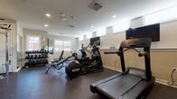 Located at 289 Ridge Rd in Wethersfield, CT, this single-floor fitness center is thoughtfully designed to accommodate a comprehensive range of workout routines. The space is organized into distinct functional zones, each equipped with professional-grade exercise equipment. The cardio area features multiple treadmills with individual monitor screens, an elliptical trainer, a stationary bike, and a rowing machine, all positioned to maximize usability and flow. A dedicated strength-training zone houses a multi-station cable machine, stability balls, and an adjustable weight bench, while a well-stocked dumbbell rack with a full-length wall mirror supports free weight training. Throughout the facility, durable rubber flooring provides both safety and impact absorption. Recessed lighting illuminates the space evenly, complemented by natural light streaming through multiple white-framed windows positioned along the walls. Neutral-toned walls and clean white wainscoting contribute to a bright, organized aesthetic. Large wall-mounted mirrors are strategically placed to enhance the sense of space and allow users to monitor their form. A sanitation station with cleaning supplies and a waste receptacle underscores the facility's attention to cleanliness and user convenience.