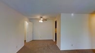 This single-floor space presents a clean, functional layout centered around a carpeted main room finished with neutral-toned walls and a textured ceiling. A ceiling fan with an integrated light fixture is centrally mounted, providing both air circulation and ambient lighting throughout the space. The room's soft, beige carpet adds warmth underfoot and contributes to the overall understated aesthetic.

The layout includes a paneled door on the left side of the room, likely leading to a closet, offering practical storage. An open passageway on the right side leads to an adjacent area, visible in the background as a darker space, suggesting a hallway, bathroom, or additional room beyond. The partial wall framing this passageway adds a subtle architectural detail to the otherwise straightforward design.

White baseboards line the perimeter of the room, providing a clean contrast against the neutral carpet and walls. The overall ambiance is simple and utilitarian, with an emphasis on practicality and ease of use. The space offers a blank canvas well-suited for personalization, making it adaptable to a variety of furnishing styles and functional needs.