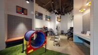 This single-floor pet grooming and activity space is designed with both functionality and fun in mind. The interior boasts soaring ceilings with exposed ductwork and industrial-style pendant lighting, lending the space an open, airy feel. White subway tile walls provide a clean backdrop, complemented by polished concrete floors throughout the main grooming areas.

The grooming zone is outfitted with multiple stainless steel grooming tables, adjustable grooming arms, and deep stainless steel wash basins equipped with handheld sprayers. A floating vanity-style sink station with gray tile accents and warm pendant lighting serves as an additional washing area. Storage units and waste bins are conveniently positioned throughout.

A dedicated play area features vibrant artificial turf flooring, a colorful tunnel crawl toy with paw print detailing, and small agility-style equipment, creating an engaging environment for pets. Colorful, dog-themed artwork and framed prints decorate the walls, adding personality and visual interest throughout the space.

A glass-paneled entry door with sidelight allows natural light to filter into the space, and small red and blue mushroom-style stools near the entrance provide casual seating. Track and pendant lighting ensures the entire space is well-illuminated.