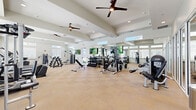 Located at 400 Hidden Palm Circle in Kissimmee, Florida, this fitness center offers a generously proportioned, single-floor workout facility designed to accommodate a wide range of exercise needs. The space features soaring ceilings with coffered architectural detailing and multiple ceiling fans that promote airflow and comfort throughout. Large windows positioned along the walls, complemented by transom windows above, flood the interior with abundant natural light while providing views of the surrounding outdoor environment.

The facility is divided into clearly defined workout zones. The cardio area is lined with a row of modern treadmills, elliptical trainers, and stationary bikes, all positioned to take advantage of the natural light streaming in from the expansive windows. The strength training section is equally well-appointed, featuring cable machines, multi-station weight systems, adjustable benches, and a comprehensive dumbbell rack spanning a wide range of weights.

Full-length mirrors are strategically placed throughout the space, enhancing the sense of openness while allowing users to monitor their form during workouts. Mounted flat-screen televisions provide entertainment throughout the facility. The light-toned carpet tile flooring ties the space together with a clean, neutral aesthetic. The overall ambiance is bright, airy, and purposefully organized, making this fitness center a well-rounded and inviting amenity.