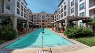 This upscale outdoor amenity space is centered around a generously sized resort-style swimming pool, framed by a classic multi-story building facade featuring arched windows, stone detailing, and private balconies. The pool area is thoughtfully designed with a shallow wading ledge lined with in-water lounge chairs, ideal for relaxation, while a decorative bronze sculpture serves as a refined focal point within the water. Surrounding the pool, a broad stone-tiled deck offers ample space for sunbathing, furnished with rows of contemporary chaise lounges. Multiple private cabanas are positioned along the pool's perimeter, each equipped with retractable curtains, ceiling fans, mounted televisions, and comfortable seating — providing shaded retreats with direct pool views. Lush landscaping, including manicured shrubs, tropical palms, and mature trees, lines the walkways and garden areas adjacent to the pool, adding a natural, resort-like ambiance. A herringbone-patterned paver walkway winds through the garden zone, featuring additional outdoor seating with navy blue cushioned furnishings. The space is accessed through the Clubhouse entrance, marked by elegant limestone block cladding and floor-to-ceiling glass double doors, setting a sophisticated tone for the entire outdoor amenity experience.