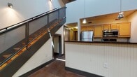 This two-story industrial loft blends raw architectural character with functional modern living. The main level is anchored by soaring double-height ceilings and polished concrete floors that lend an urban, warehouse-inspired aesthetic. A standout feature is the large glass block window set within an exposed brick wall, flooding the living area with diffused natural light while maintaining privacy. The open-concept kitchen is well-appointed with warm wood cabinetry, dark granite countertops, stainless steel appliances including a refrigerator and microwave, and a breakfast bar with beadboard paneling. A dramatic metal-railed staircase with wood-tread steps connects the main level to the upper loft, which features light hardwood flooring, exposed concrete ceiling beams, and brick accent walls. The upper level also benefits from a skylight, enhancing the airy, open feel. A dedicated closet area with wire shelving and multiple paneled doors provides ample storage. The space includes two full bathrooms, each with a tub-shower combination, tile flooring, and wood cabinetry. Throughout, dark baseboards and gray-toned doors provide cohesive finishing touches that complement the loft's industrial character.