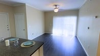 This single-floor space offers an open and functional layout connecting the kitchen and living areas. The living room is defined by dark hardwood flooring that contrasts cleanly with the neutral, light beige walls, creating a warm yet understated ambiance. Crown molding runs along the ceiling perimeter, adding a refined architectural detail to the space. A ceiling fan with a built-in light fixture is centrally positioned, providing both air circulation and ambient lighting.

A standout feature of the living area is the large sliding glass door dressed with vertical blinds, which allows an abundance of natural light to pour into the room. This element also suggests access to an outdoor area, further enhancing the sense of openness. Multiple electrical outlets are distributed along the walls, offering practical utility throughout the space.

Adjacent to the living area, the kitchen is partially visible and features dark granite countertops, complementing the flooring tones and adding a touch of sophistication. The overall design is clean, cohesive, and move-in ready, appealing to those seeking a straightforward and well-maintained living environment.