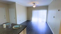 This single-floor space offers an open and functional layout connecting the kitchen and living areas. The living room is defined by dark hardwood flooring that contrasts cleanly with the neutral, light beige walls, creating a warm yet understated ambiance. Crown molding runs along the ceiling perimeter, adding a refined architectural detail to the space. A ceiling fan with a built-in light fixture is centrally positioned, providing both air circulation and ambient lighting.

A standout feature of the living area is the large sliding glass door dressed with vertical blinds, which allows an abundance of natural light to pour into the room. This element also suggests access to an outdoor area, further enhancing the sense of openness. Multiple electrical outlets are distributed along the walls, offering practical utility throughout the space.

Adjacent to the living area, the kitchen is partially visible and features dark granite countertops, complementing the flooring tones and adding a touch of sophistication. The overall design is clean, cohesive, and move-in ready, appealing to those seeking a straightforward and well-maintained living environment.