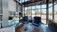 Located on Cadiz Street in Dallas, Texas, this single-floor space offers a thoughtfully designed interior that blends industrial and contemporary elements throughout. The entrance is defined by dramatic floor-to-ceiling steel-framed glass walls that flood the space with natural light while framing direct views of the Dallas downtown skyline.

The main lounge area features rich hardwood flooring, built-in shelving, and generous seating arrangements, including deep sofas and high-back upholstered chairs positioned to take full advantage of the expansive city views. Decorative planters line the interior window sills, adding a natural element to the modern aesthetic.

A dedicated coffee and dining area — branded as a brew station — incorporates reclaimed wood accent walls, exposed wood beam ceiling details, pendant lighting clusters, and a large communal table surrounded by cobalt blue velvet chairs. This area is equipped with a full beverage station, creating a functional and visually distinctive gathering space.

A glass-enclosed conference room provides a more formal setting, featuring a wood-paneled ceiling, textured brick accent walls, a large oval conference table, and an elegant multi-bulb chandelier. The space is accessible through full-height glass sliding doors, maintaining visual openness while offering privacy.