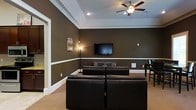 This single-floor space centers around a well-appointed lounge or media room designed for comfort and entertainment. The room features rich, dark olive-brown walls accented by crisp white crown molding and a bold white chair rail that runs the perimeter, creating a classic two-tone effect. A tray ceiling with recessed lighting adds architectural depth overhead, while a ceiling fan with a built-in light fixture ensures both comfort and ambient illumination. Wall-to-wall neutral carpeting contributes to the room's warm, inviting atmosphere. The focal point of the space is a wall-mounted flat-screen television, positioned at an ideal viewing height and flanked by a tall floor lamp that provides soft supplemental lighting. Black leather seating is arranged to face the screen, offering a dedicated media-viewing setup. A piece of wall art adds a touch of personality to the space. An adjacent area is partially visible through a doorway, hinting at additional rooms beyond. The overall ambiance is sophisticated yet comfortable, with a cohesive dark-and-white color palette that lends a modern, polished character throughout.