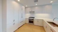 Located on the 19th floor at 8800 Lombard in San Diego, this residence delivers a striking modern aesthetic defined by clean lines, an all-white palette, and an abundance of natural light. Light-toned hardwood flooring runs throughout the living areas and bedrooms, complementing the crisp white walls and creating a bright, airy atmosphere.

The open living area is anchored by expansive floor-to-ceiling windows and glass sliding doors that frame sweeping panoramic views of the San Diego cityscape, lush treetops, and distant mountain ranges. A private balcony extends the living space outdoors, further showcasing the elevated vantage point.

The kitchen features flat-front cabinetry in a soft grey finish, quartz countertops, a marble-style backsplash, stainless steel appliances including a gas range and oven, and integrated refrigerator panels — all illuminated by recessed lighting.

The bathroom is finished with large-format marble-look tile flooring, a soaking tub with tiled surround, a floating vanity with quartz countertop, undermount sink, and a backlit mirror that adds a refined, contemporary touch. A secondary bathroom offers a bright, all-white design with a large window framing open views. The spacious walk-in closet provides ample built-in shelving and hanging rods for generous storage.