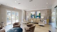 Located at 2910 Clairmont Rd in Atlanta, GA, this single-floor community lounge space offers a refined and welcoming environment designed for both relaxation and social interaction. The interior is finished with light hardwood flooring, crisp white crown molding, and recessed lighting throughout, lending a polished, contemporary aesthetic.

The main lounge area is anchored by a comfortable neutral-toned sofa and an assortment of upholstered accent chairs arranged around a glass-top coffee table, all set atop a natural jute area rug. Bold, oversized abstract artworks in teal and green tones serve as a striking focal point on the textured feature wall, while flanking table lamps add warm ambient lighting. Large glass-paneled doors flood the space with natural light and open directly onto a landscaped outdoor patio, seamlessly connecting the interior to lush greenery.

A secondary seating nook near floor-to-ceiling windows features a high-top table with bar-height chairs and pendant globe lighting, offering a more intimate setting with garden views. A glass-walled private room with a coffered ceiling provides a semi-enclosed area visible from the main lounge, adding functional versatility. The overall ambiance is sophisticated yet approachable, characterized by soft neutral tones, curated décor, and an abundance of natural light.