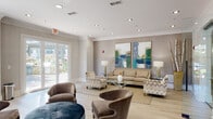 Located at 2910 Clairmont Rd in Atlanta, GA, this single-floor community lounge space offers a refined and welcoming environment designed for both relaxation and social interaction. The interior is finished with light hardwood flooring, crisp white crown molding, and recessed lighting throughout, lending a polished, contemporary aesthetic.

The main lounge area is anchored by a comfortable neutral-toned sofa and an assortment of upholstered accent chairs arranged around a glass-top coffee table, all set atop a natural jute area rug. Bold, oversized abstract artworks in teal and green tones serve as a striking focal point on the textured feature wall, while flanking table lamps add warm ambient lighting. Large glass-paneled doors flood the space with natural light and open directly onto a landscaped outdoor patio, seamlessly connecting the interior to lush greenery.

A secondary seating nook near floor-to-ceiling windows features a high-top table with bar-height chairs and pendant globe lighting, offering a more intimate setting with garden views. A glass-walled private room with a coffered ceiling provides a semi-enclosed area visible from the main lounge, adding functional versatility. The overall ambiance is sophisticated yet approachable, characterized by soft neutral tones, curated décor, and an abundance of natural light.