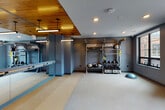 This single-floor fitness studio is thoughtfully designed to support a wide range of workout styles in a sleek, contemporary environment. The space is defined by a striking design contrast: deep slate-blue walls pair with a warm, natural wood-paneled ceiling inset that anchors the mirror wall area, adding visual warmth to the otherwise cool, modern aesthetic.

A full-length wall of floor-to-ceiling mirrors runs along one side of the studio, complete with a mounted ballet barre — ideal for stretching, barre workouts, and functional movement training. Overhead, pendant linear lighting fixtures hang from the wood ceiling panel, providing focused illumination across the open exercise floor.

The workout area is equipped with a RaX functional training rig, offering TRX suspension straps alongside neatly organized shelving stocked with medicine balls, kettlebells, and a BOSU balance trainer — providing a comprehensive range of strength and conditioning tools. The flooring transitions between large-format tile near the entrance and a cushioned, speckled rubber surface across the main workout zone, supporting both high-impact and low-impact activities.

Natural light filters in through black-framed windows, offering views to the outdoors and keeping the space feeling open and connected to its surroundings. A dedicated mechanical access door is discreetly integrated into the layout. Overall, the studio delivers a professional, motivating atmosphere well-suited for functional fitness and group or individual training.