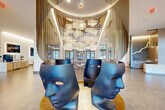 Located at 903 Monroe St SW in Huntsville, Alabama, this expansive single-floor amenity and lobby space is a showcase of contemporary design and artistic vision. The centerpiece of the space is a dramatic rotunda-style focal point featuring cascading metallic chain curtains, oversized cloud-like pendant lighting, and sculptural face-shaped chairs that command immediate attention upon entry. Soaring ceilings and floor-to-ceiling black-framed glass windows flood the interiors with abundant natural light while offering views of the surrounding outdoor spaces and courtyard. Flooring transitions between large-format tile and herringbone wood patterns, adding visual depth and warmth throughout. A staffed reception area with a sleek white desk anchors one end of the space, while curated seating vignettes — including curved sofas, lounge chairs, and colorful geometric rugs — create distinct social zones. A decorative laser-cut metal screen partition adds an organic, nature-inspired element, complementing the organic sculptural art pieces. Additional amenities include a shuffleboard table, a built-in bookcase wall with circular porthole accents, and a large wall-mounted display with backlit framing. The overall ambiance is bold, modern, and gallery-like, blending art-forward design with functional communal living.