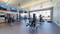 This modern fitness center is thoughtfully designed to support a comprehensive workout experience. The space is anchored by large floor-to-ceiling windows along one wall, flooding the interior with abundant natural light and offering open views to the outside. The layout is spacious and organized, with dedicated zones for both strength training and cardiovascular exercise. Strength training equipment includes cable machines, weight benches, and various resistance machines, while the cardio area is outfitted with treadmills, ellipticals, stair climbers, and stationary bikes. The flooring features durable, dark rubberized material suited for high-traffic fitness use. A full-length mirrored wall, framed in a bold orange accent, expands the sense of space and allows users to monitor their form. Multiple ceiling fans are suspended throughout, promoting airflow and comfort during intense workouts. Several large flat-screen TVs are mounted on the walls, providing entertainment and added motivation. Overhead linear lighting ensures the space remains bright and well-lit at all hours. The overall ambiance is clean, modern, and functional, creating an inviting environment for fitness enthusiasts of all levels.