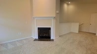 This single-floor space presents a clean, understated aesthetic defined by neutral beige walls and plush wall-to-wall carpeting that lends a sense of warmth and comfort. The living area centers around a classic fireplace framed by a crisp white mantel with traditional millwork detailing, set against a floor-to-ceiling chimney surround. The firebox features decorative logs, adding a cozy, functional element to the room. A dark stone or tile hearth anchors the fireplace and provides a grounded contrast to the light carpeting surrounding it. White chair rail molding runs along the lower portion of the walls, adding a subtle architectural detail that complements the clean trim work throughout. Recessed lighting in the ceiling provides controlled ambient illumination, contributing to the room's tidy, well-maintained appearance. The overall palette — warm beige walls, white trim, and light carpet — creates a cohesive, neutral canvas that would easily accommodate a variety of interior design styles. The space feels open and move-in ready, with no clutter or furnishings to distract from its architectural features.