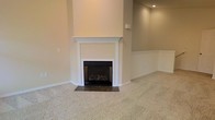 This single-floor space presents a clean, understated aesthetic defined by neutral beige walls and plush wall-to-wall carpeting that lends a sense of warmth and comfort. The living area centers around a classic fireplace framed by a crisp white mantel with traditional millwork detailing, set against a floor-to-ceiling chimney surround. The firebox features decorative logs, adding a cozy, functional element to the room. A dark stone or tile hearth anchors the fireplace and provides a grounded contrast to the light carpeting surrounding it. White chair rail molding runs along the lower portion of the walls, adding a subtle architectural detail that complements the clean trim work throughout. Recessed lighting in the ceiling provides controlled ambient illumination, contributing to the room's tidy, well-maintained appearance. The overall palette — warm beige walls, white trim, and light carpet — creates a cohesive, neutral canvas that would easily accommodate a variety of interior design styles. The space feels open and move-in ready, with no clutter or furnishings to distract from its architectural features.