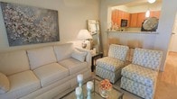 This single-floor residence offers a thoughtfully designed layout that balances comfort and functionality. The living area serves as the heart of the home, furnished with a plush cream sofa, patterned accent chairs, and a glass-top coffee table that lend the space a light, airy feel. Decorative touches such as pillar candles, fresh floral arrangements, and a large botanical wall art piece add character and warmth to the room. A full-length mirror and a classic table lamp contribute to the bright, welcoming ambiance. The living space flows seamlessly into the kitchen through an open-concept pass-through, defined by a raised counter with bar seating — ideal for casual dining or entertaining. The kitchen features rich orange-toned cabinetry, light stone countertops, and modern appliances including a built-in microwave, creating a vibrant contrast to the soft neutral palette of the living area. Neutral wall tones and soft carpeting throughout the living space tie the design together, resulting in a cohesive and inviting environment. The overall aesthetic is clean, contemporary, and carefully curated, making this a comfortable and visually appealing home.