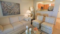 This single-floor residence offers a thoughtfully designed layout that balances comfort and functionality. The living area serves as the heart of the home, furnished with a plush cream sofa, patterned accent chairs, and a glass-top coffee table that lend the space a light, airy feel. Decorative touches such as pillar candles, fresh floral arrangements, and a large botanical wall art piece add character and warmth to the room. A full-length mirror and a classic table lamp contribute to the bright, welcoming ambiance. The living space flows seamlessly into the kitchen through an open-concept pass-through, defined by a raised counter with bar seating — ideal for casual dining or entertaining. The kitchen features rich orange-toned cabinetry, light stone countertops, and modern appliances including a built-in microwave, creating a vibrant contrast to the soft neutral palette of the living area. Neutral wall tones and soft carpeting throughout the living space tie the design together, resulting in a cohesive and inviting environment. The overall aesthetic is clean, contemporary, and carefully curated, making this a comfortable and visually appealing home.