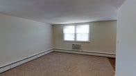 This single-floor space presents a straightforward and functional layout designed with practicality in mind. The main room is finished with wall-to-wall carpeting in a neutral beige tone, providing a warm underfoot feel that complements the light-colored walls throughout. A pair of windows along one wall allows natural light to filter into the room, creating a bright and airy atmosphere while offering a view of surrounding greenery outside.

Climate control is well-considered, with a wall-mounted air conditioning unit positioned beneath the windows for efficient cooling during warmer months. Baseboard heating runs along the perimeter of the room, ensuring consistent warmth during cooler seasons. The combination of both systems provides year-round comfort.

The walls are finished in a clean, off-white tone that enhances the sense of openness within the room. The ceiling is smooth and uniform, contributing to the overall clean aesthetic of the space. The room's generous proportions offer ample flexibility for a variety of furniture arrangements and uses. Overall, the space provides a neutral, move-in-ready environment suited for comfortable everyday living.