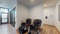 This thoughtfully designed single-floor spa offers a serene and contemporary environment tailored for relaxation and wellness. The space features warm-toned luxury vinyl plank flooring throughout, lending a clean and cohesive aesthetic that complements the bright, white walls and recessed lighting overhead.

A dedicated relaxation nook houses two full-body black leather massage chairs, positioned side by side against crisp white walls. This area is equipped with a wall-mounted control panel and ventilation, providing a quiet, private retreat for guests. Glass sliding doors partially enclose the space, allowing natural light to filter in while maintaining a sense of intimacy.

The main corridor connects the various areas of the spa and features a striking accent wall finished in a warm gold tone with an artistic drip detail, adding a bold visual element to the otherwise neutral palette. Built-in white cabinetry with gold hardware lines one wall, offering ample storage, while open shelving displays books, plants, and decorative objects, contributing to a welcoming, curated atmosphere.

Framed artwork, including a standout nature-inspired print, adds personality and character to the walls. Large black-framed glass doors and windows along one side of the corridor flood the space with natural light and provide views of the surrounding exterior. The overall design balances modern minimalism with warm, inviting touches, creating a polished spa environment.